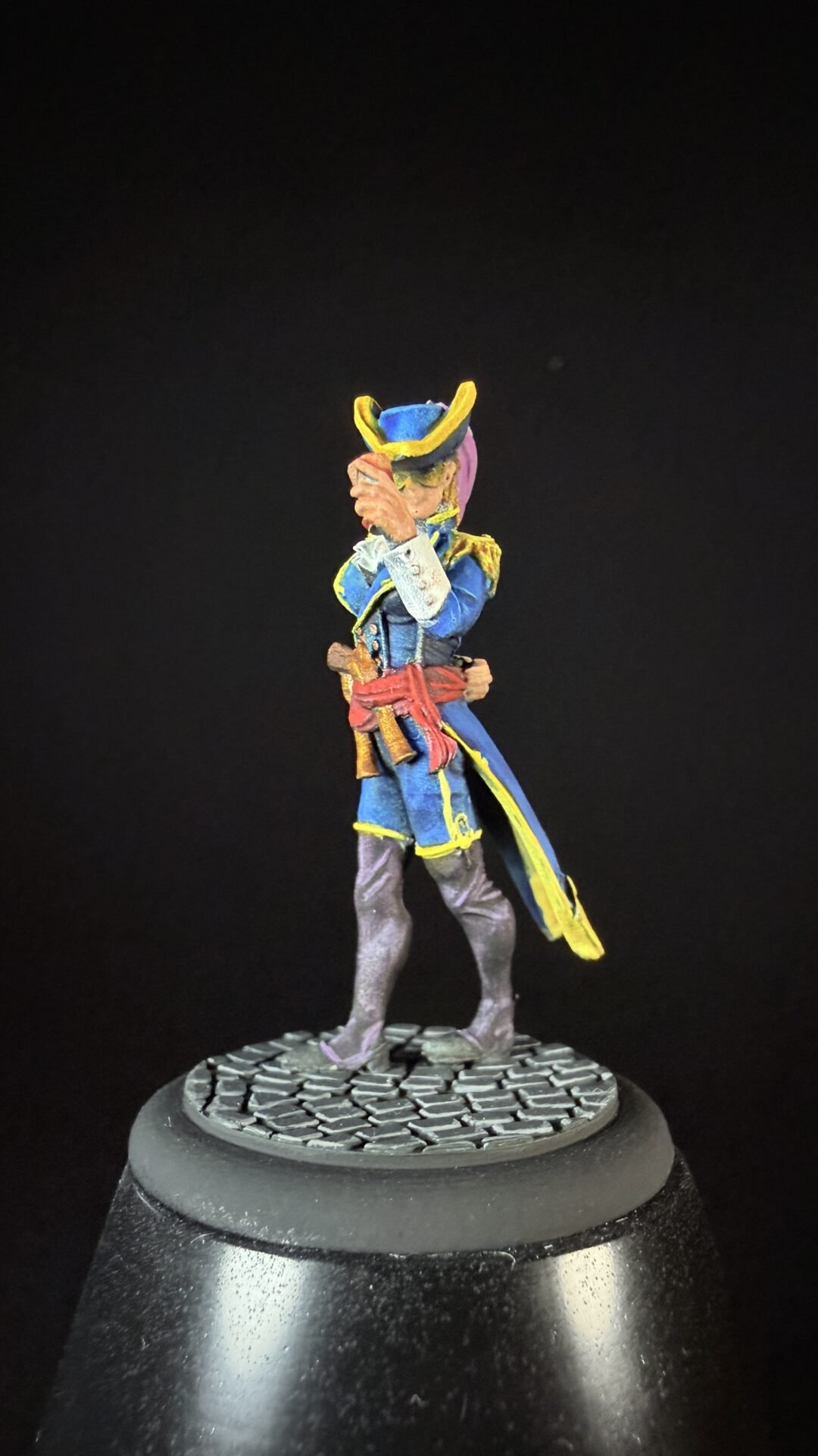 Small painted pirate miniature in a blue coat with yellow trim, tricorn hat, and red sash, standing on a textured circular base.
