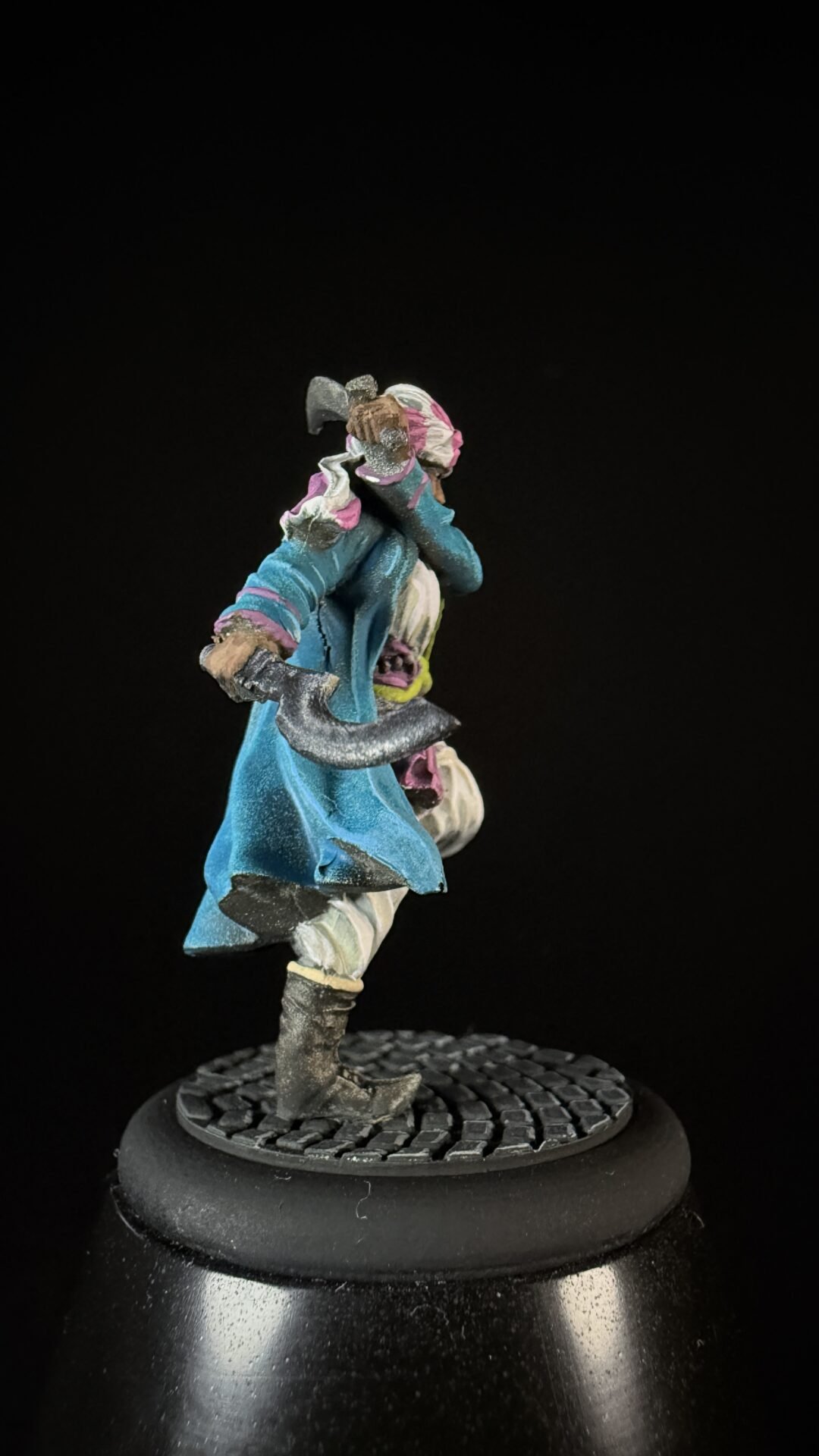 Painted miniature figure in a teal coat with pink and white scarf, wielding a curved sword, standing on a textured round base against a black background.