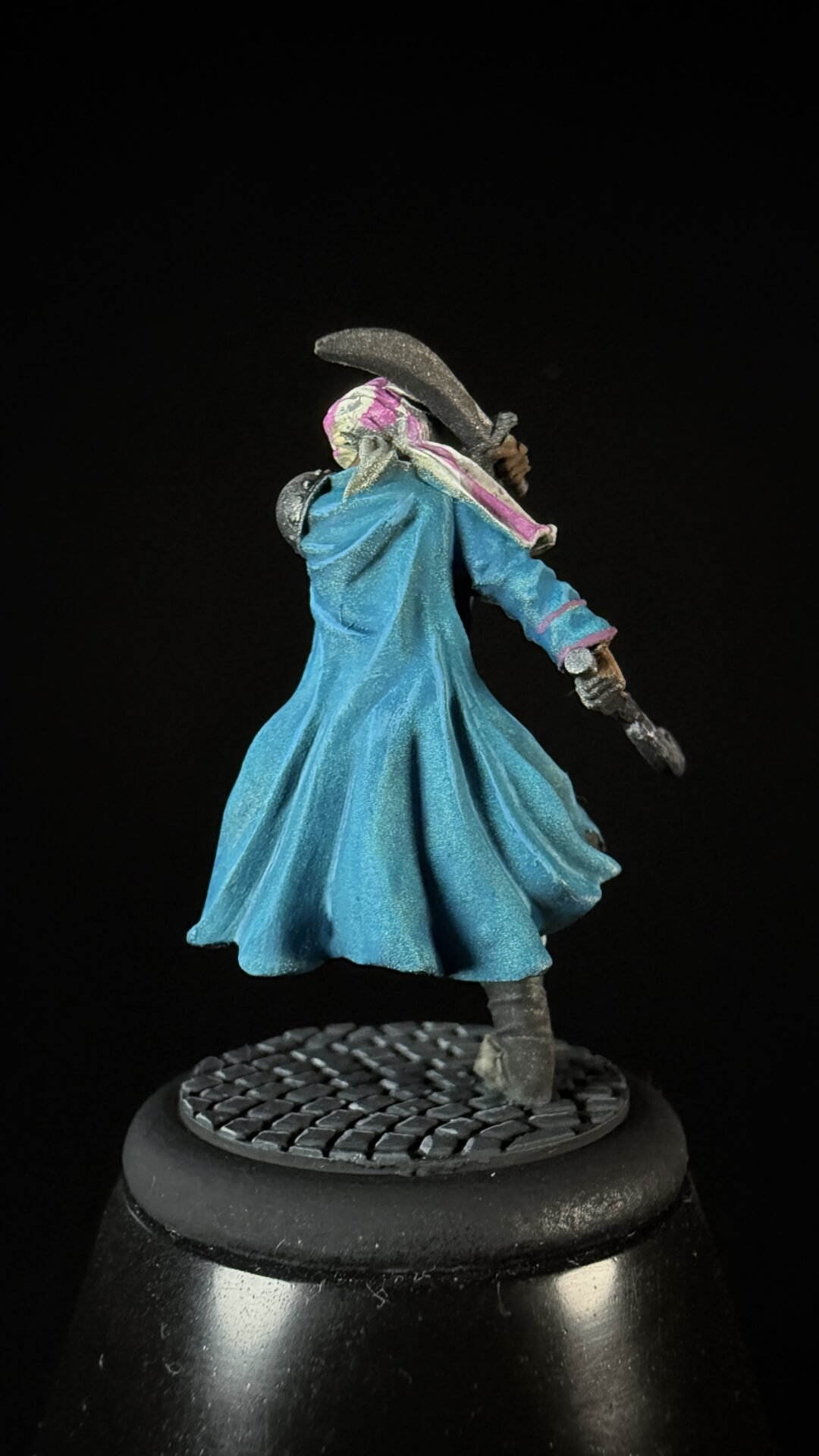 Miniature figure of a hooded traveler in a blue cloak, wielding a curved blade, posed on a textured base against a black background.