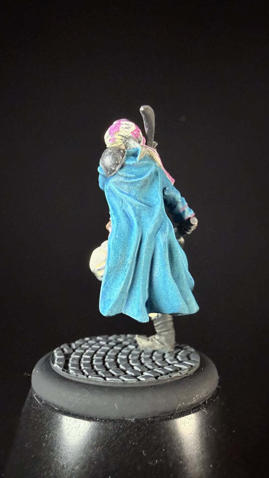 Back view of a fantasy miniature in a bright blue cloak and pink hood, standing on a textured round base with a staff over the shoulder.