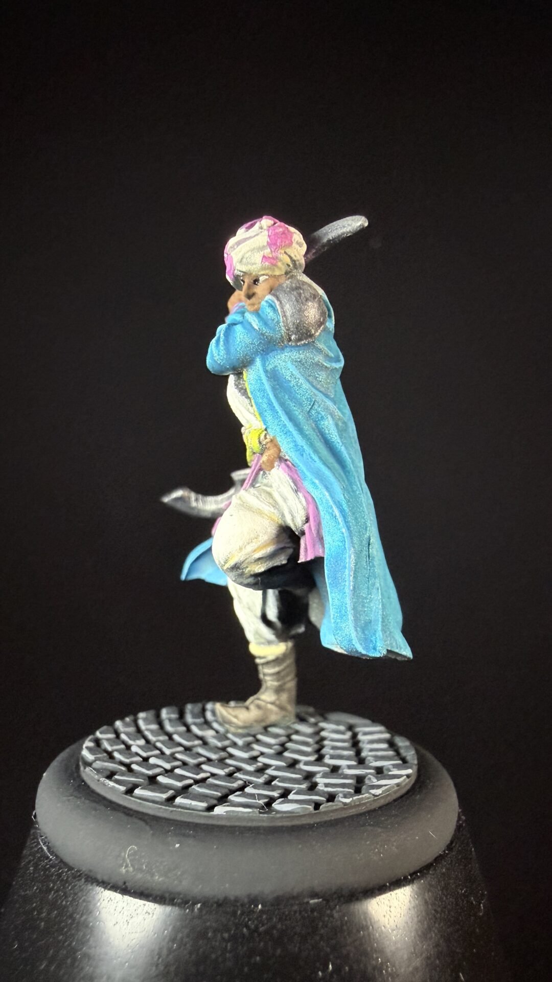 Miniature figure of a robed man in a blue cloak and pink turban, standing on a circular textured base.