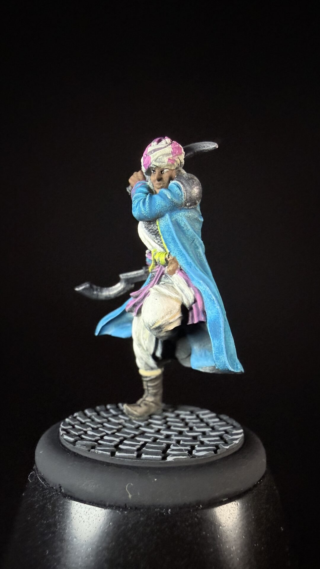 painted fantasy miniature figure wearing a turquoise-blue coat and pink turban, wielding curved blades, standing on a textured circular base on a black background