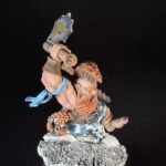 Painted tabletop dwarf miniature raising a gray hammer, with orange beard and blue scarf, standing on a rocky base.