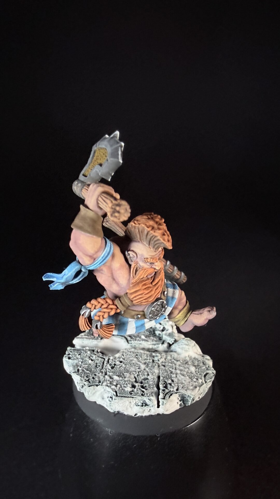 Painted tabletop dwarf miniature raising a gray hammer, with orange beard and blue scarf, standing on a rocky base.