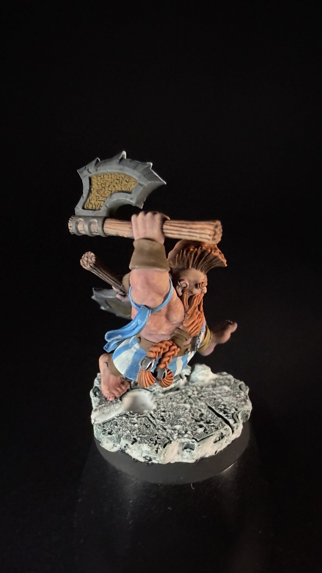 Fantasy miniature barbarian warrior swung over his head with a large double-headed axe, standing on a rocky base, wearing blue fabric and brown leather.