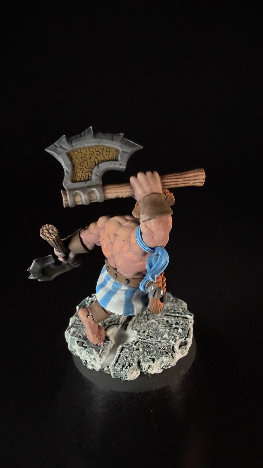 Barbarian mini figure mid-stride, lifting a large ornate axe over his shoulder, wearing a blue-and-white striped kilt and brown accessories on a rocky base.