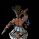 Fantasy miniature: muscular barbarian warrior with a large, rune-inscribed axe, blue scarf, and blue-and-white striped kilt, standing on a rocky base.