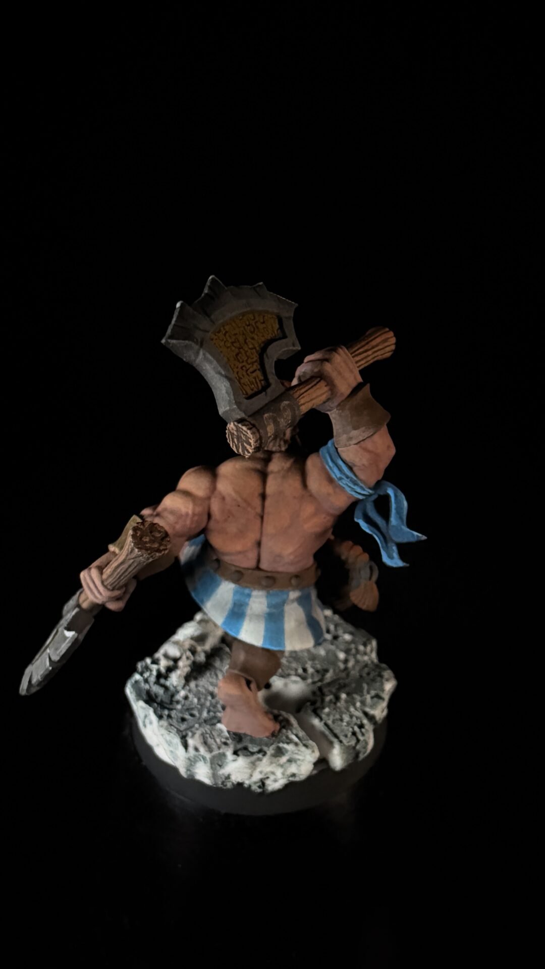 Fantasy miniature: muscular barbarian warrior with a large, rune-inscribed axe, blue scarf, and blue-and-white striped kilt, standing on a rocky base.
