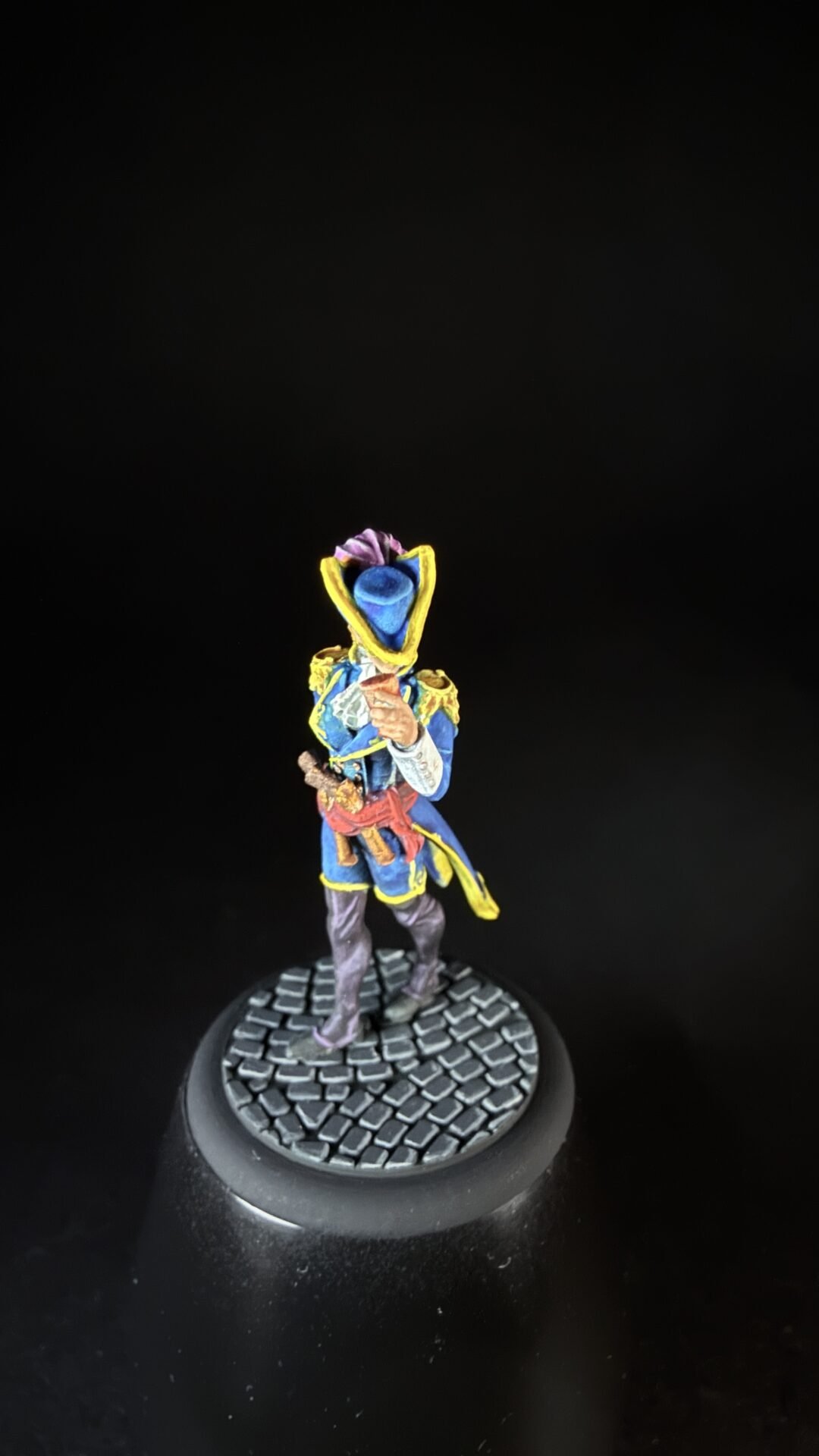 Colorful fantasy miniature: a blue-coated figure with yellow trim and purple plume, standing on a cobblestone base.