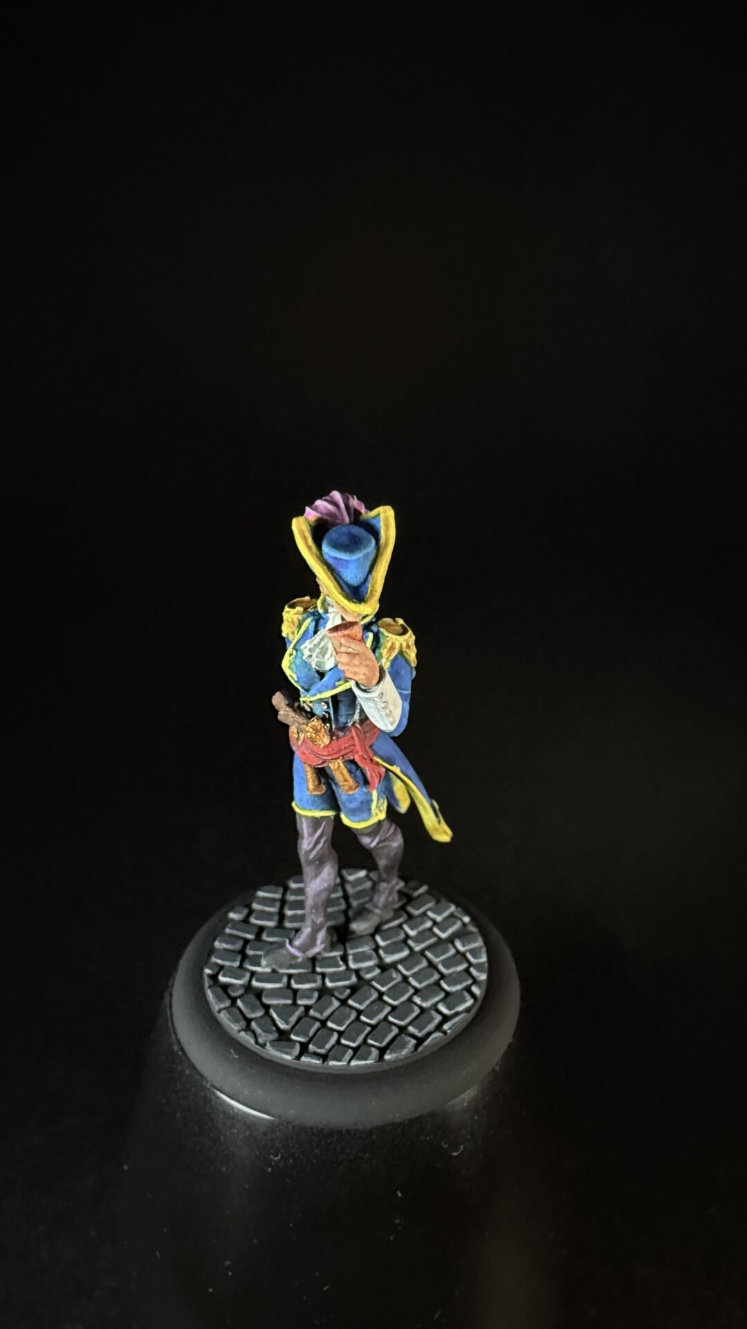 Small painted fantasy miniature figure in a blue and yellow military-style coat with a red sash, standing on a cobblestone base; the figure wears a blue helmet/face mask and holds a weapon.