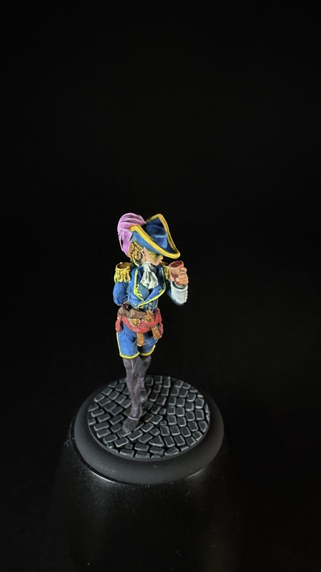 Colorful pirate miniature in a blue coat with a pink feathered hat, standing on a textured circular base and holding a pistol.