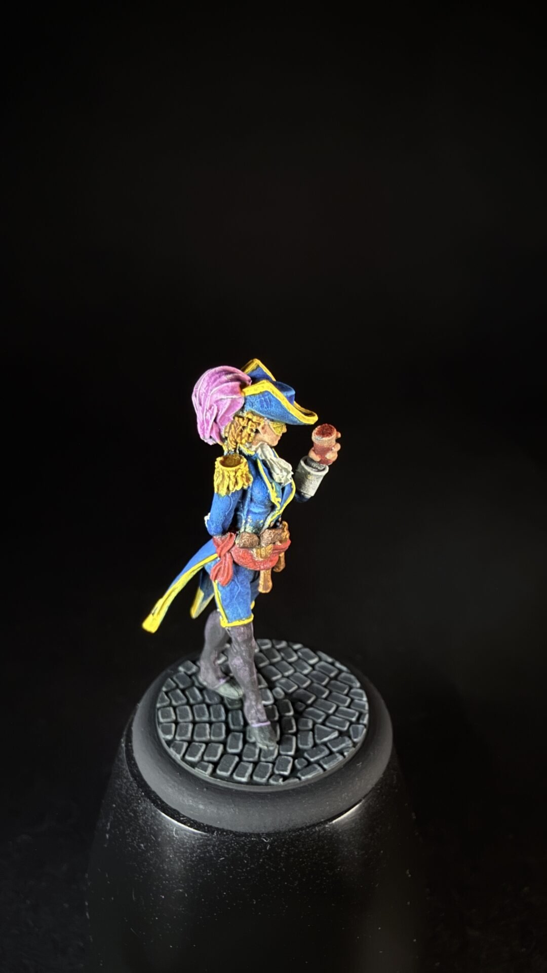 Colorful pirate miniature in a blue coat with yellow trim, holding a red object, standing on a textured circular base.