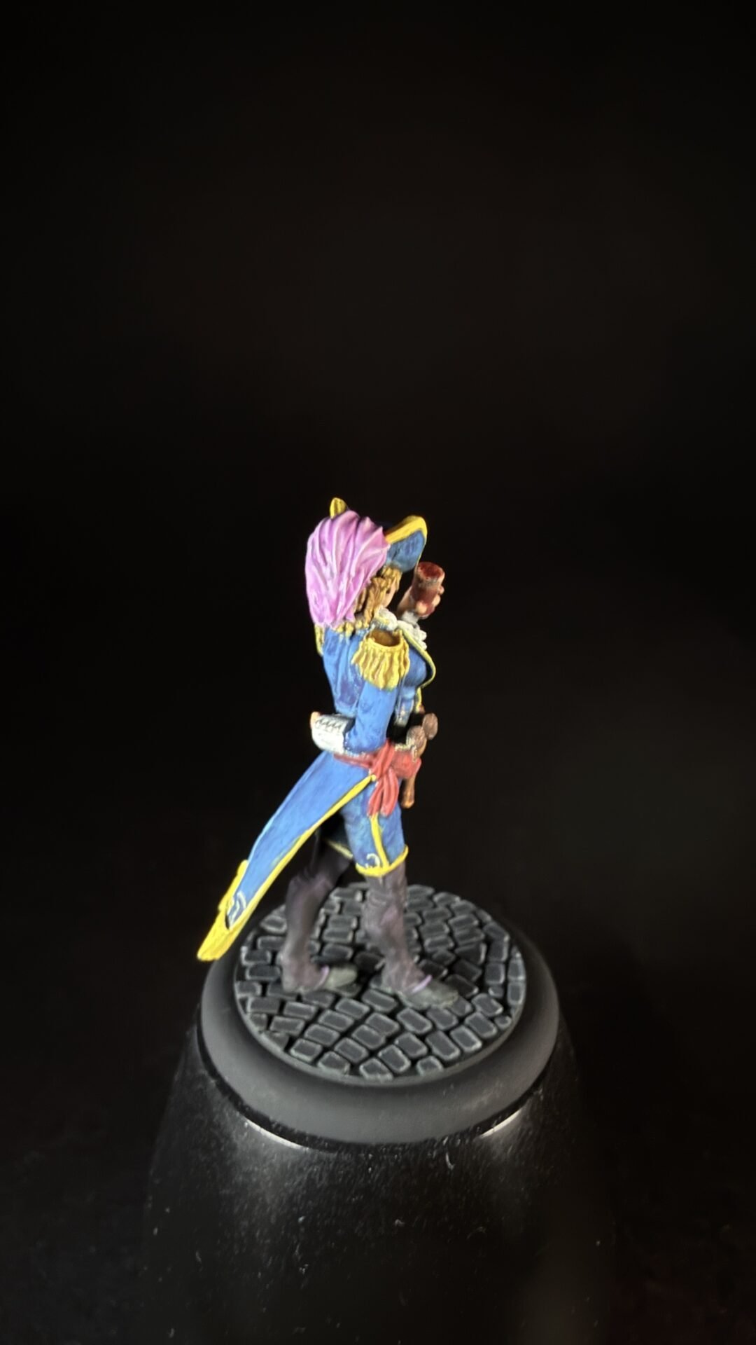 Colorful painted fantasy miniature figure in a blue coat with yellow trim and a pink plume, standing on a round cobblestone base.