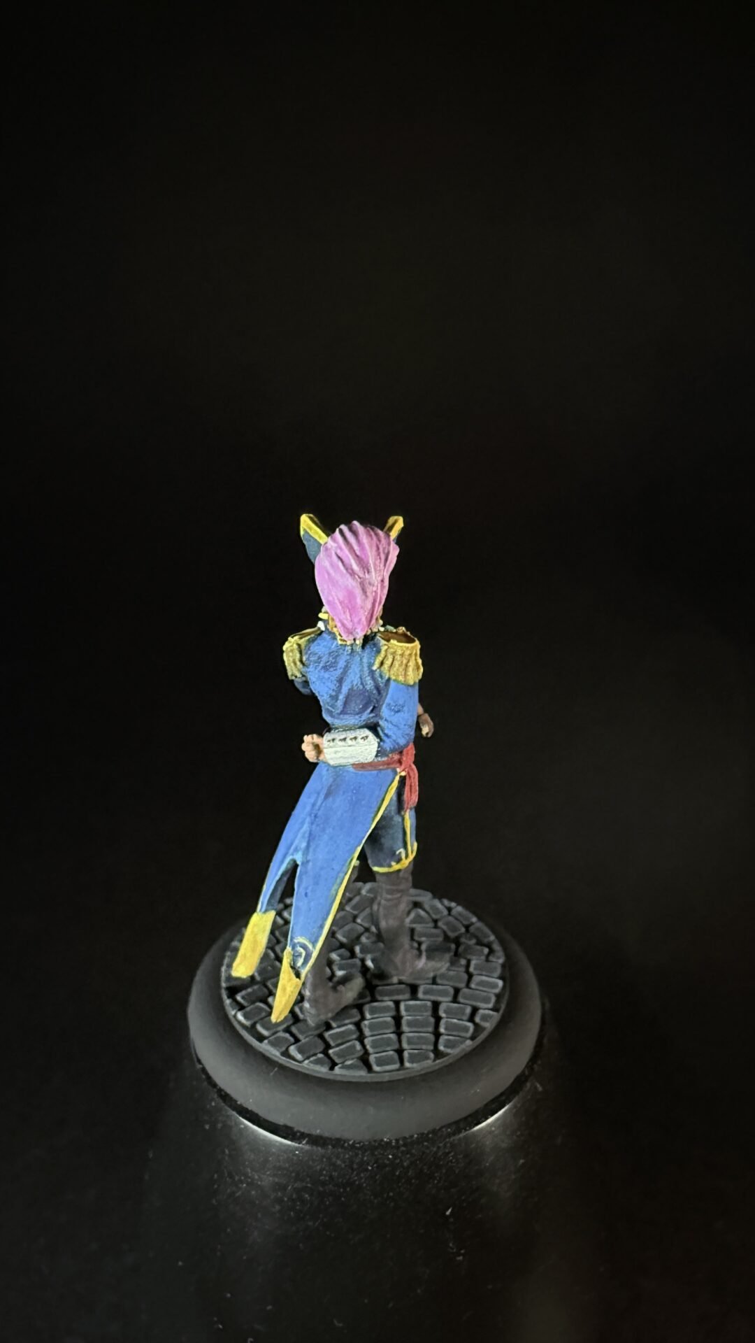 Small painted miniature figure in a blue coat with yellow epaulettes and pink headscarf, standing on a cobblestone pedestal.