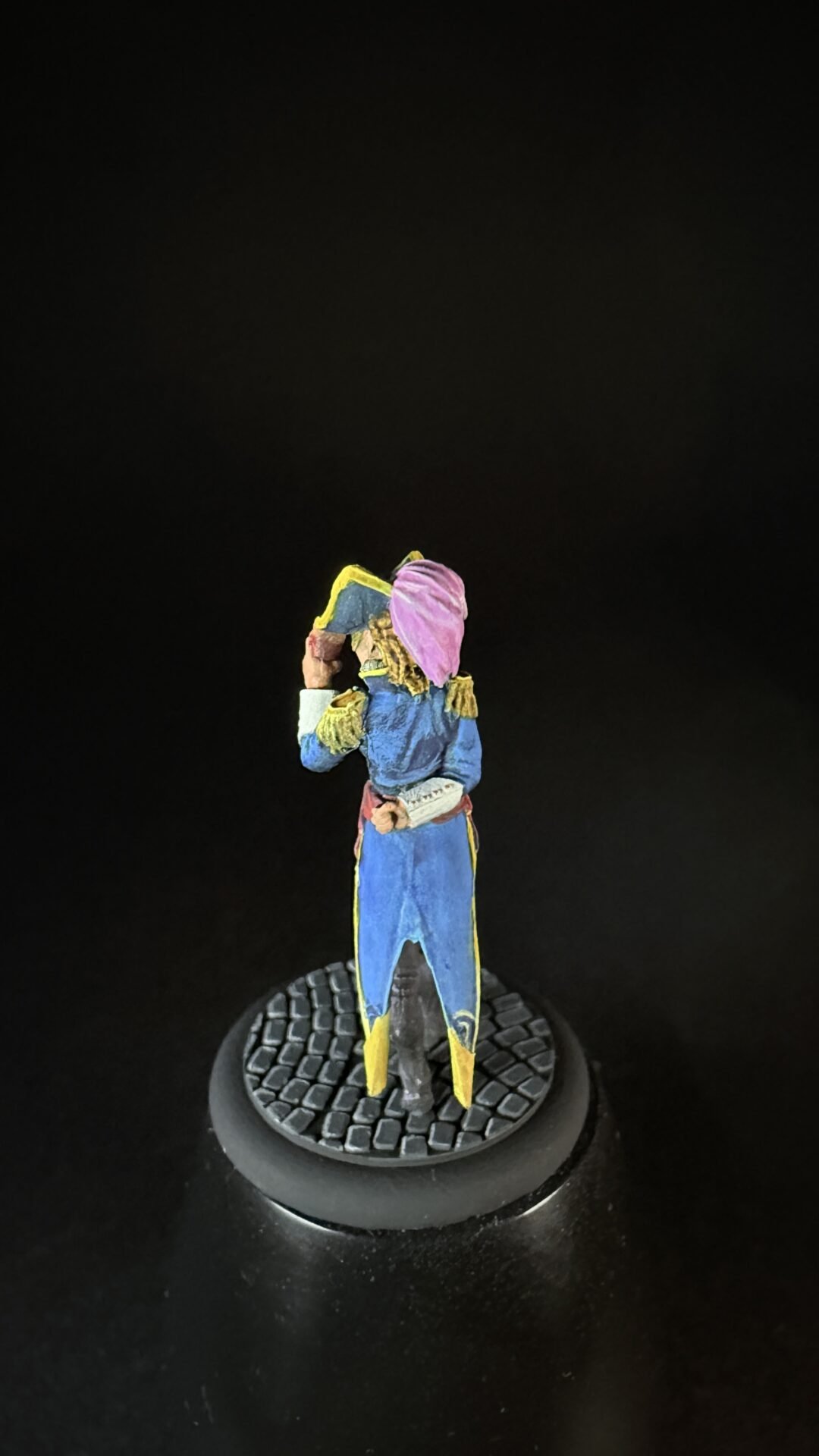 Tiny painted figurine of a person in a blue coat with yellow trim and a pink hood, standing on a round textured black base.