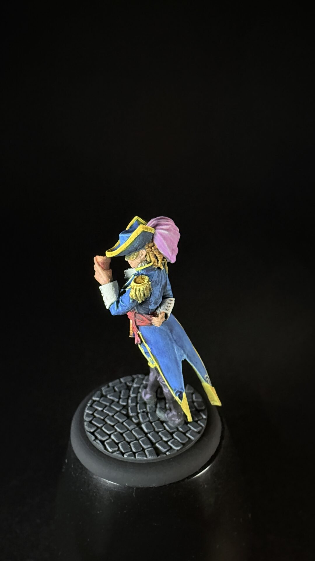Painted miniature of a Napoleonic-era officer in a blue coat with yellow trim and pink cloak, standing on a textured round base against a black background.