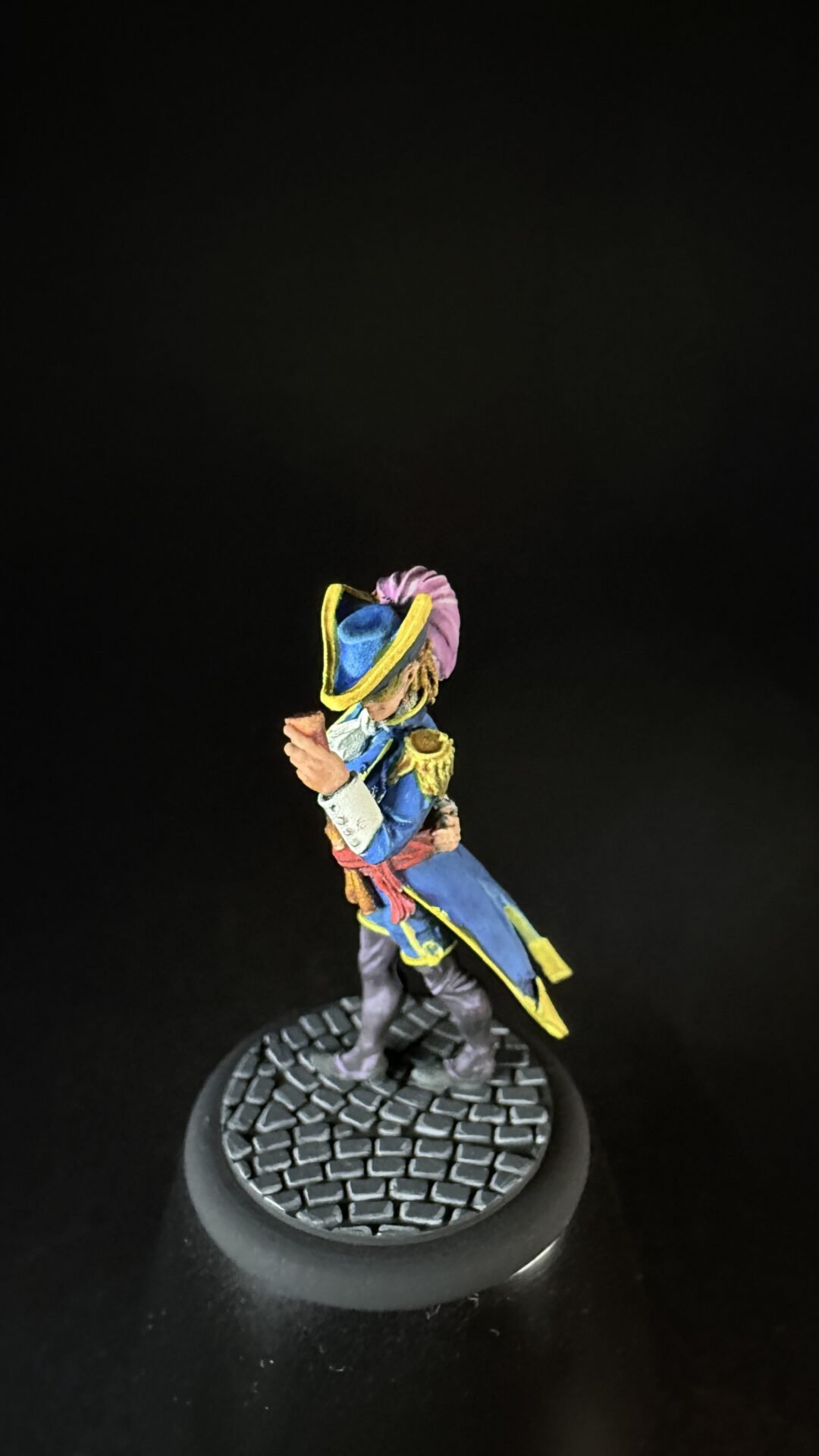 Detailed miniature figure in a blue coat with yellow trim, pink sash, and tricorn hat, standing on a textured round base against a black background.