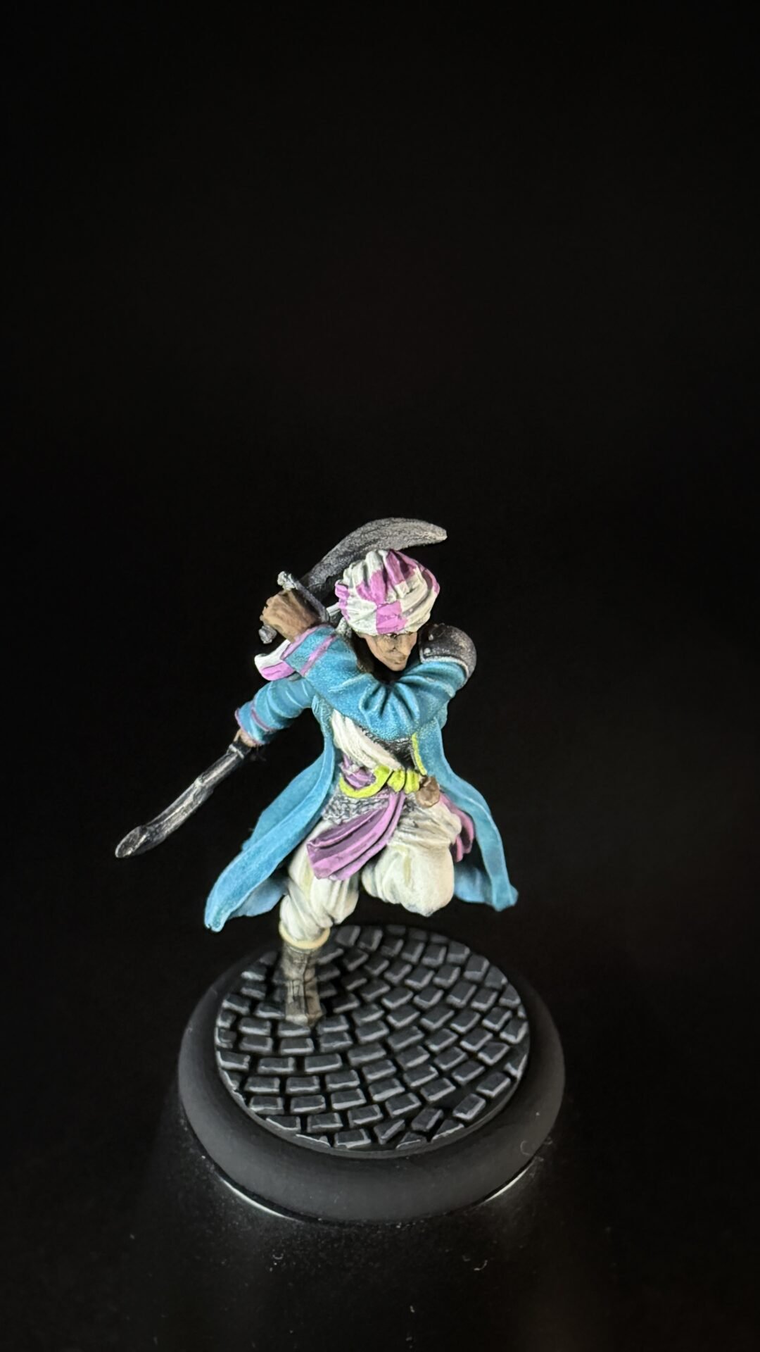 Painted miniature of a warrior in a teal blue coat with a pink headwrap, swinging a curved blade, standing on a textured round base against a black background.