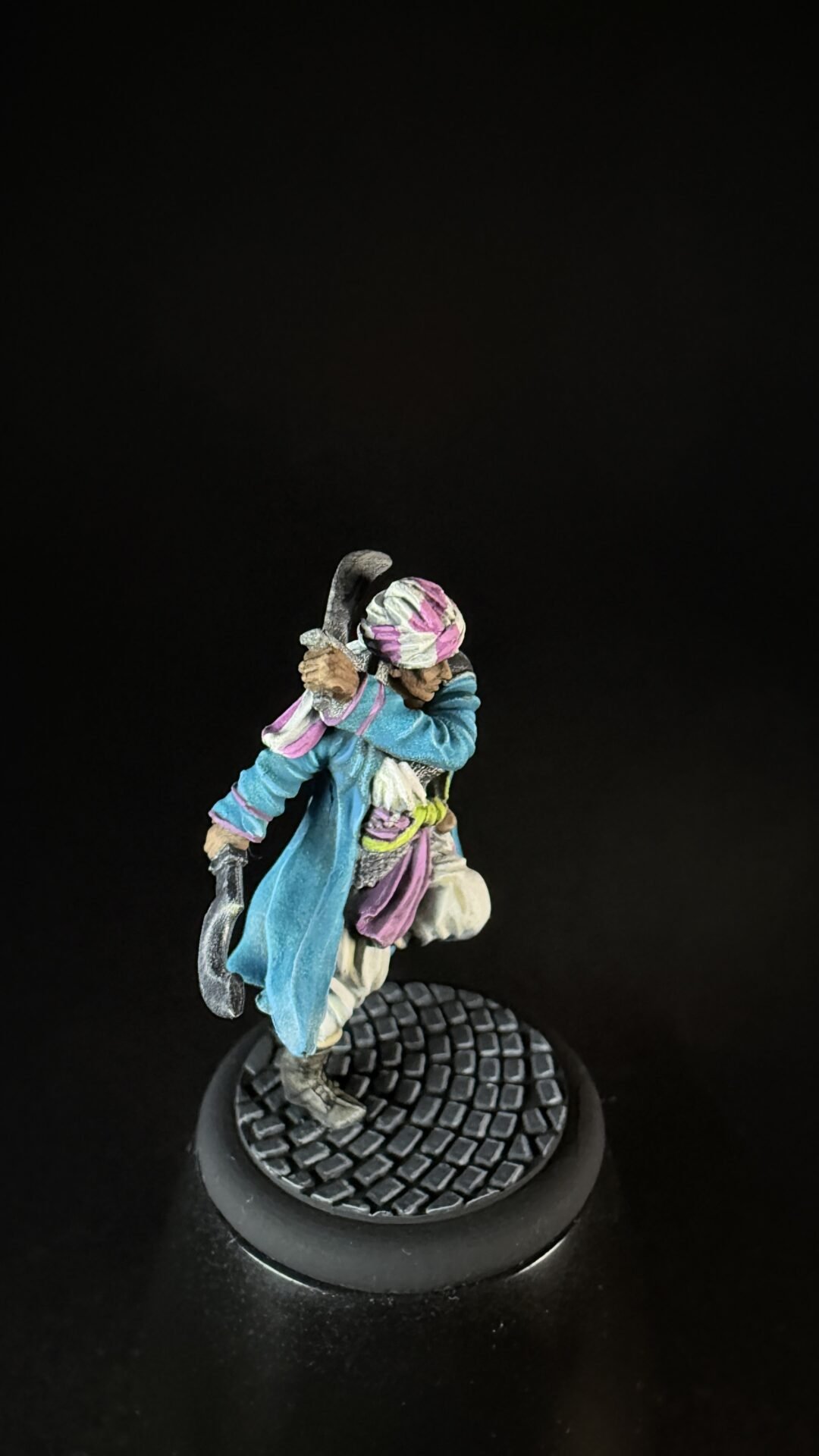 Colorful fantasy miniature figure swinging a curved sword, wearing a blue coat and pink headscarf, on a textured circular base.