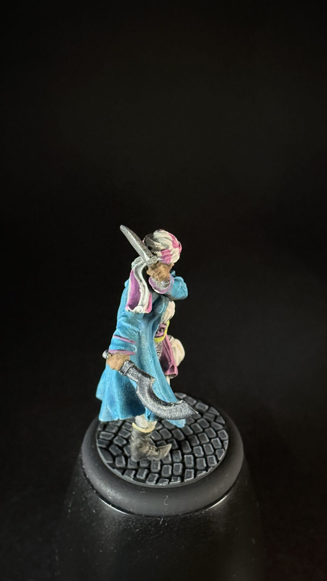 Colorful fantasy miniature: a figure in a blue cloak with pink accents, raised curved sword, standing on a textured cobblestone base against a dark background