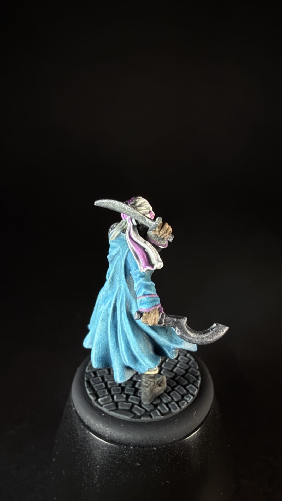 Painted tabletop miniature of a hooded fantasy figure in a blue robe wielding two curved silver blades, standing on a cobblestone base with purple accents on the scarf.