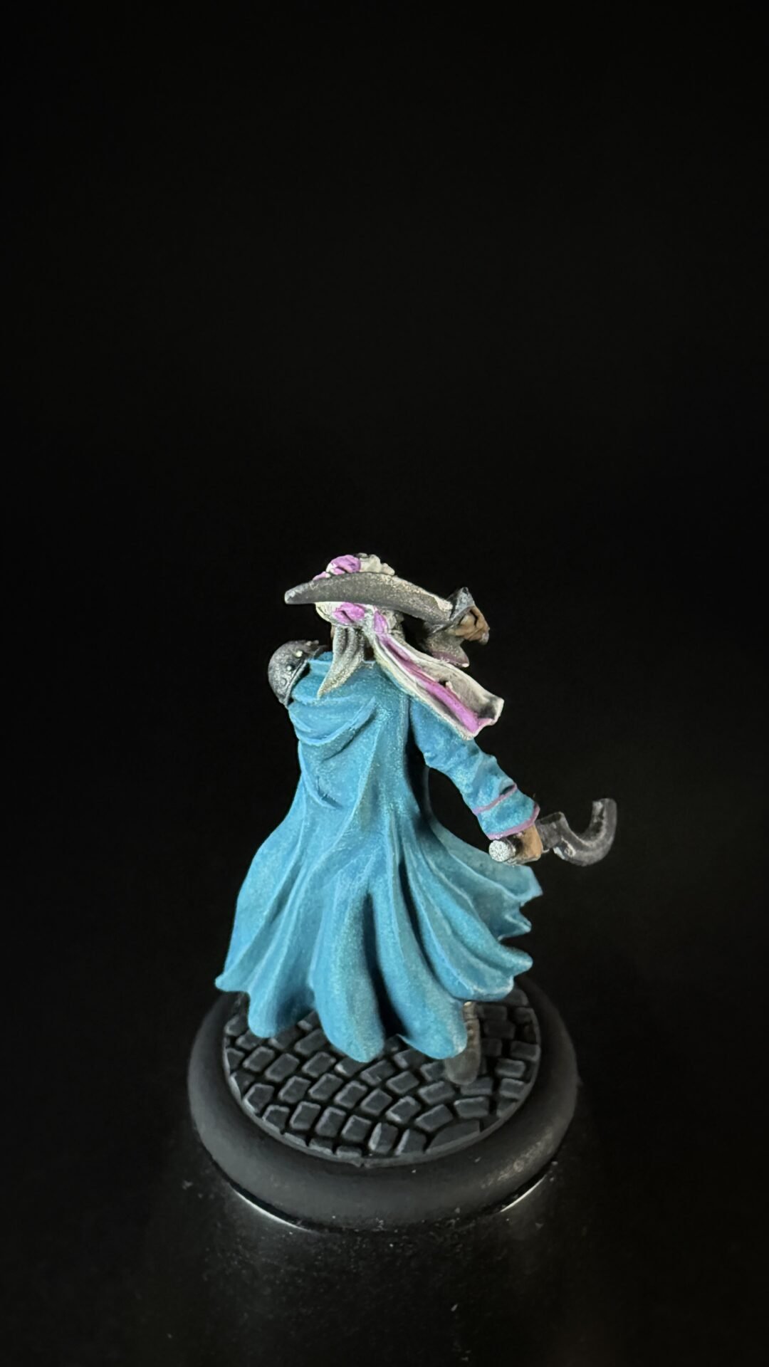 Small tabletop fantasy miniature: hooded figure in blue cloak with a pink scarf, wielding a curved blade on a textured round base.