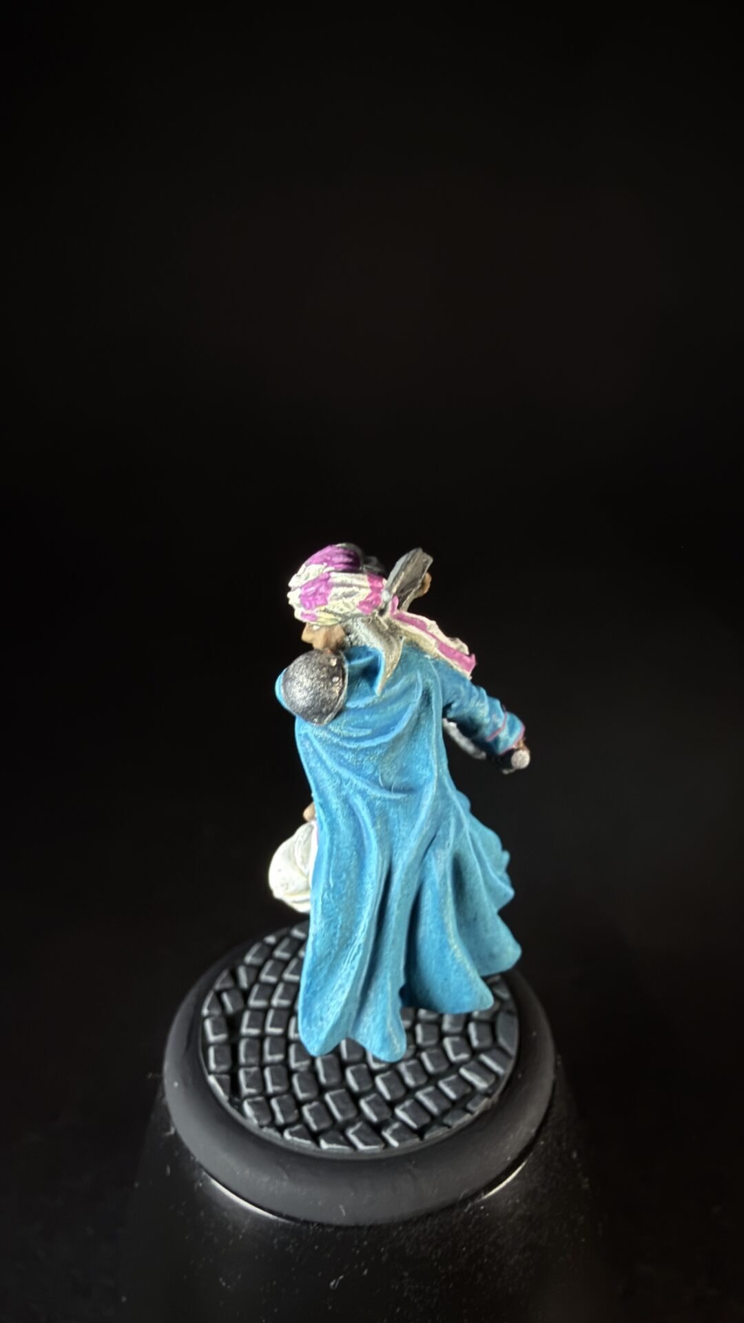 Small figurine of a person in a flowing blue cloak with a pink and white headscarf, standing on a round textured black base.