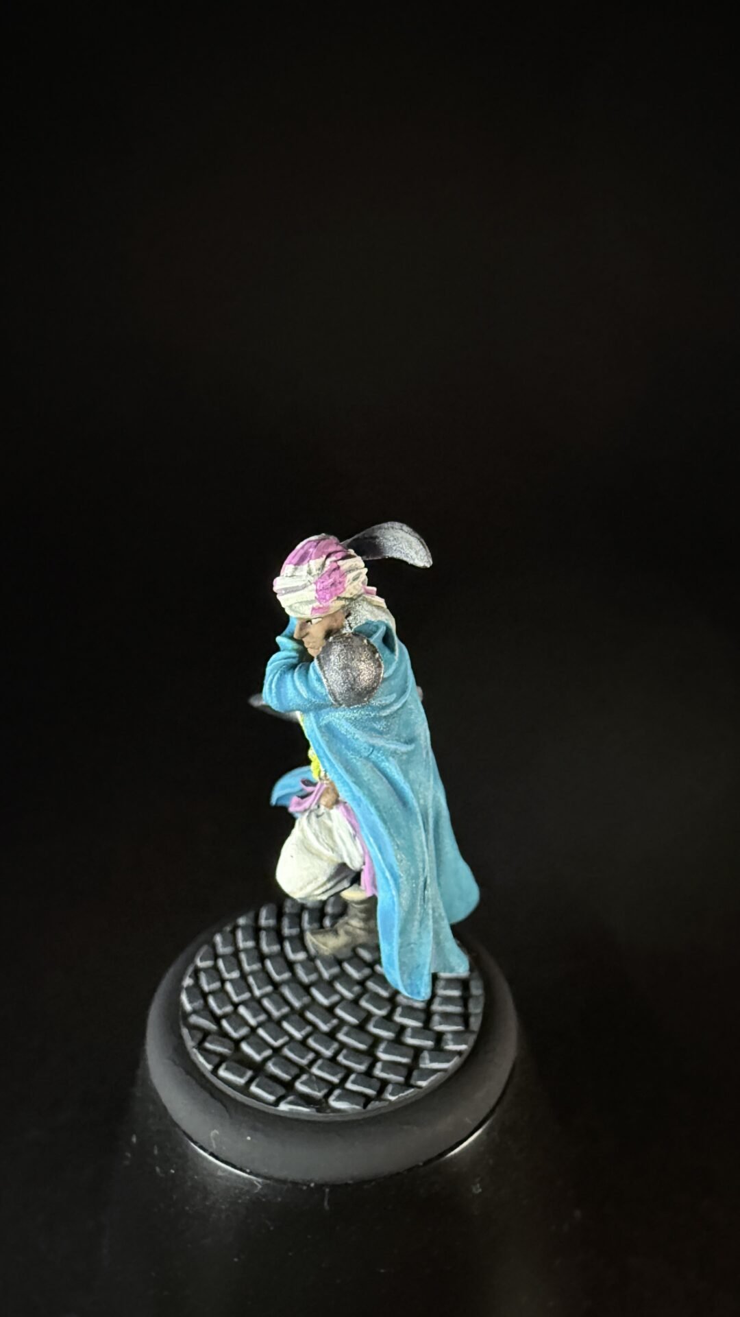 Small fantasy figurine with a blue cloak and pink headwrap on a textured display base against a dark background, seen from the side.