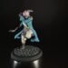 Tabletop miniature of a pirate-like fighter in a blue coat, pink scarf, and white pants, posed with a curved blade, standing on a cobblestone base against a dark background