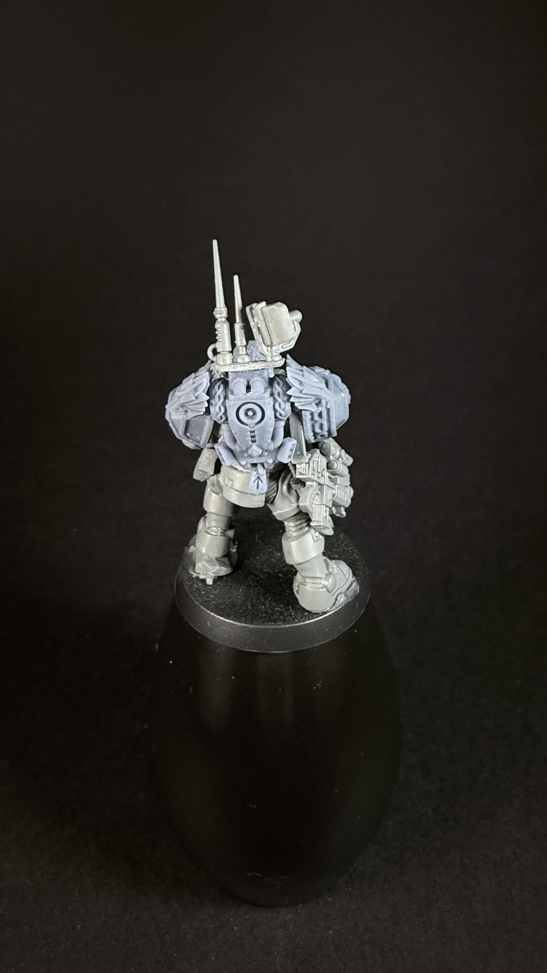 Back view of a gray armored sci-fi mech miniature with antennas and a round display base on a black background