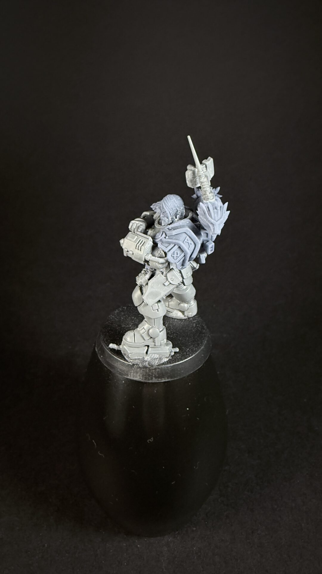 Grayscale sci‑fi armored miniature with a raised sword, standing on a round display base with gear-packed backpack and detailed plating.