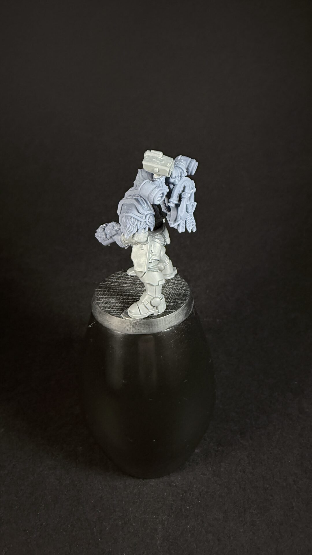 Unpainted gray sci‑fi armored miniature on a round display base, facing left, with large backpack gear and plated armor.