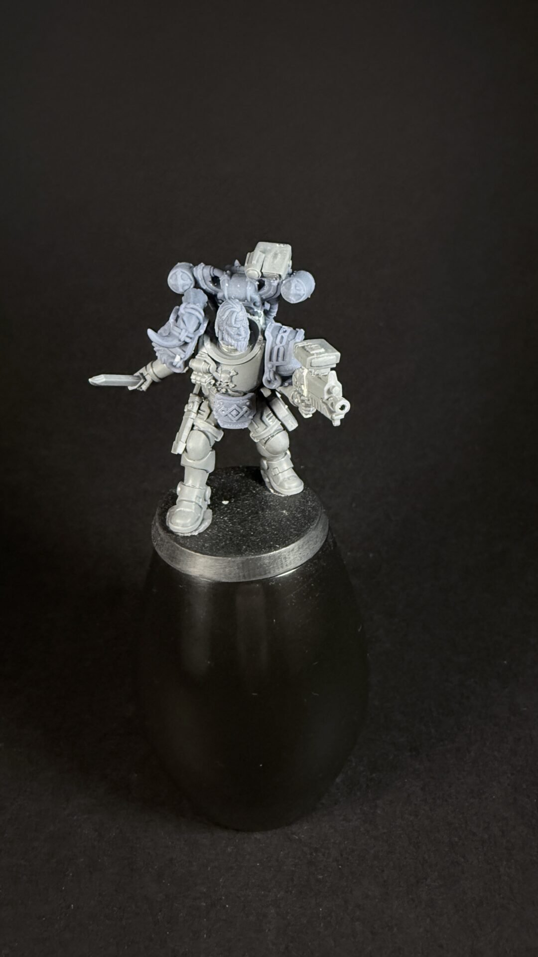 Unpainted sci‑fi miniature of a space marine in heavy power armor, holding a blade in one hand and a bolter in the other, standing on a round display base.
