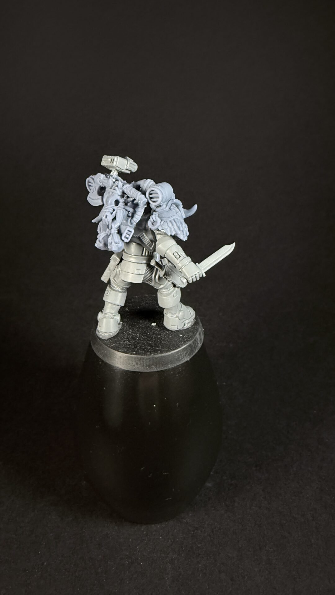 Unpainted gray sci-fi armored miniature holding a curved blade, with a bulky backpack and gear on a round base.