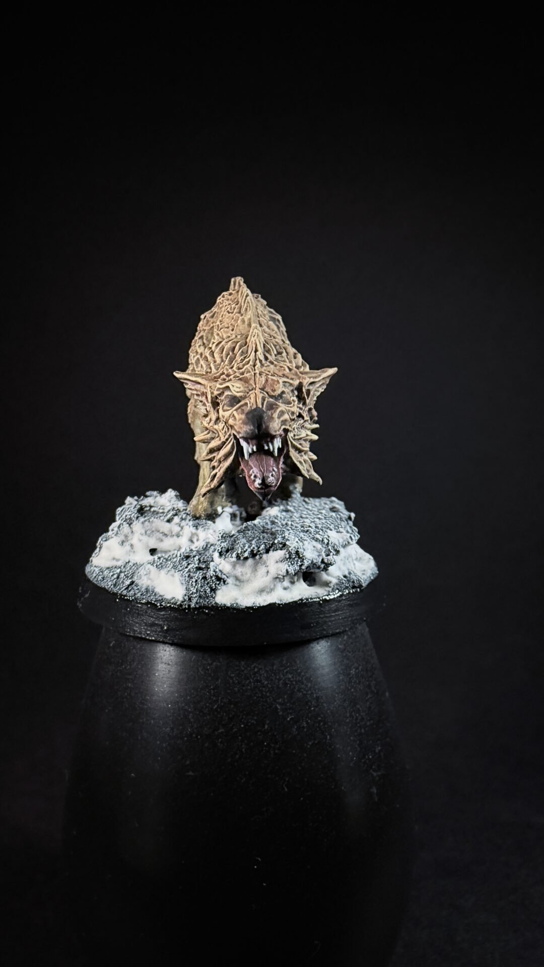 Close-up of a fantasy dragon head miniature with jagged, bark-like scales and open jaws, mounted on a snow-dusted rocky base on a black pedestal.