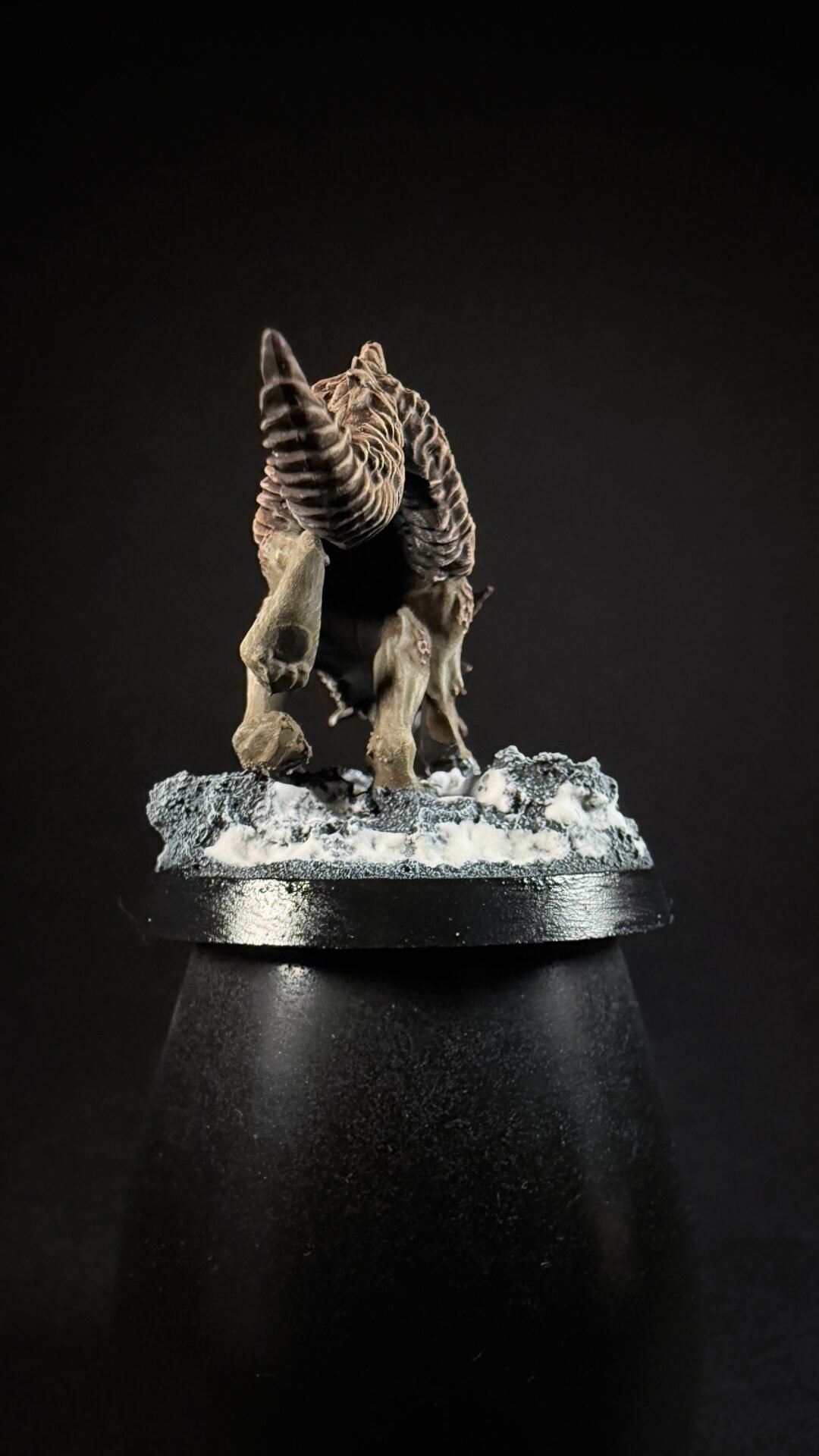 Fantasy ram-like creature with oversized spiraling horns standing on a snow-dusted rocky base against a dark background