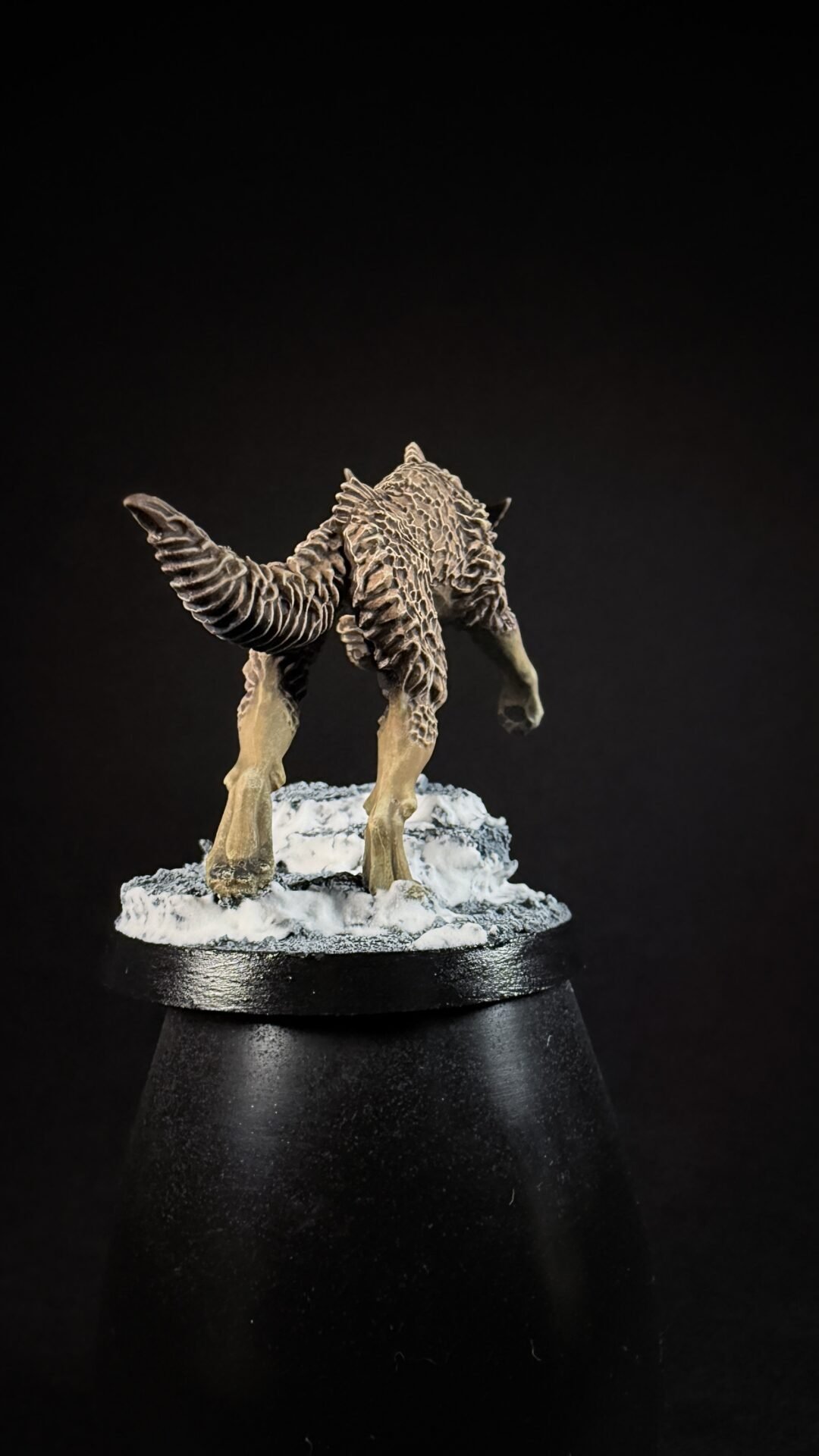 Fantasy creature mini on a snow-covered base, viewed from behind, with textured, shaggy fur and a curved tail.