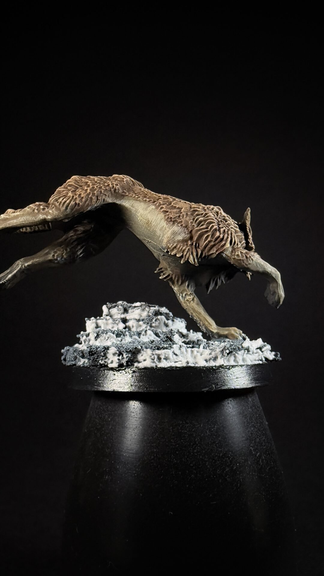 Feathered bipedal dinosaur sculpture mid-stride on a snow-covered rock base, displayed on a black pedestal against a dark background.