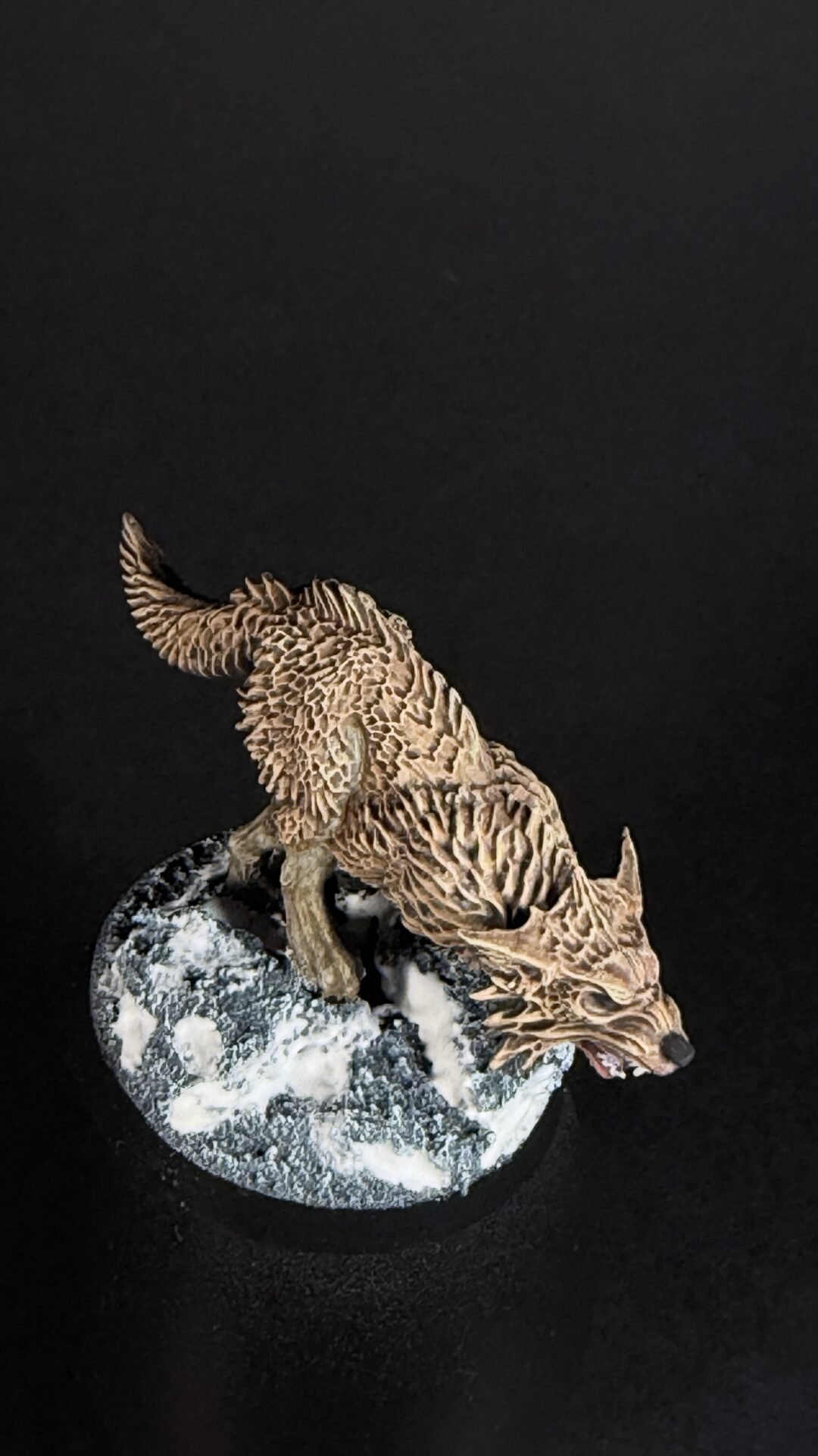 Brown dragon figurine with scaly, textured wings perched on a snow-covered rock against a black background, viewed from above.