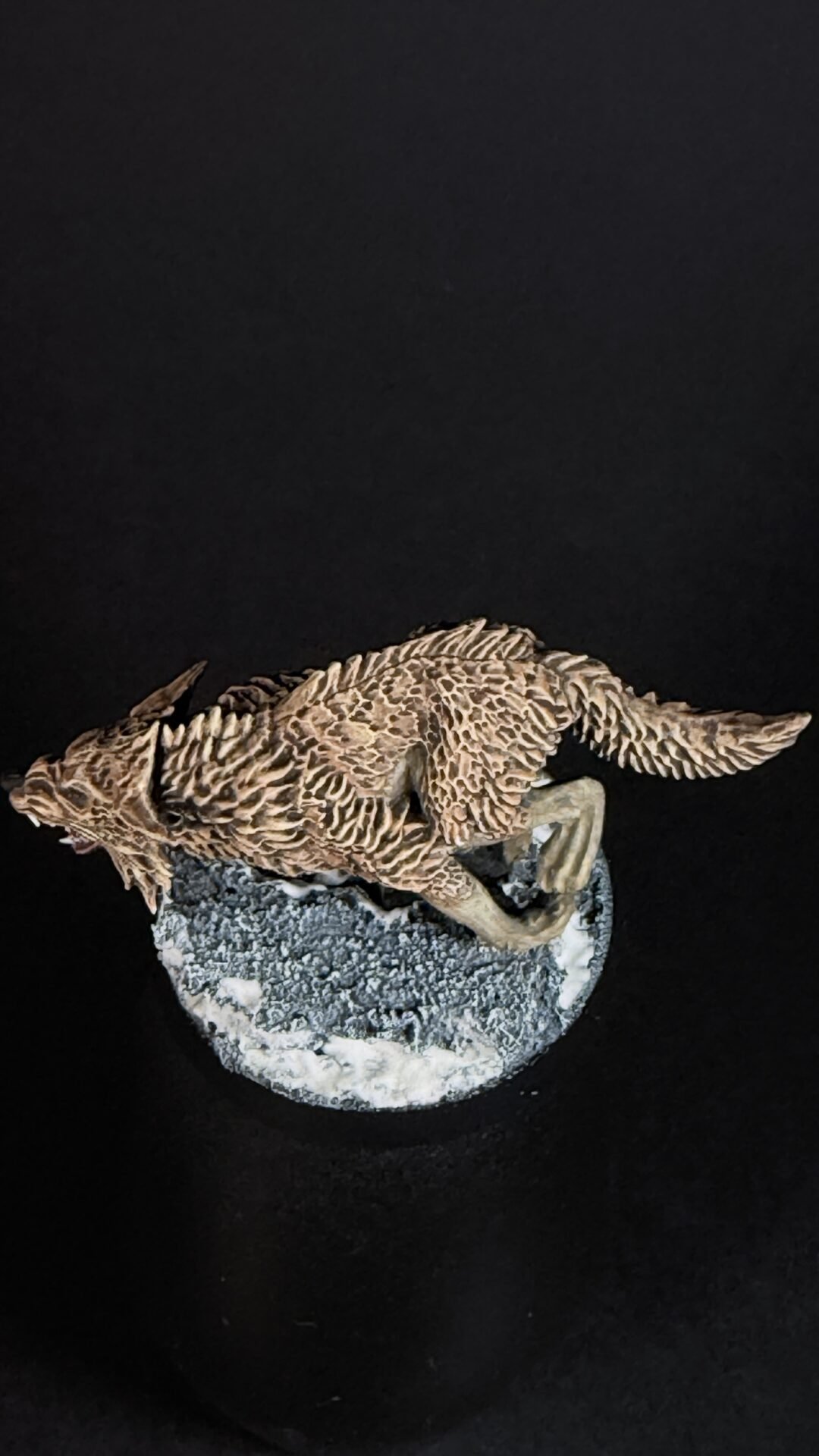 Brown textured dinosaur-like sculpture with a long tail, perched on a rocky, snow-dusted base against a black background.