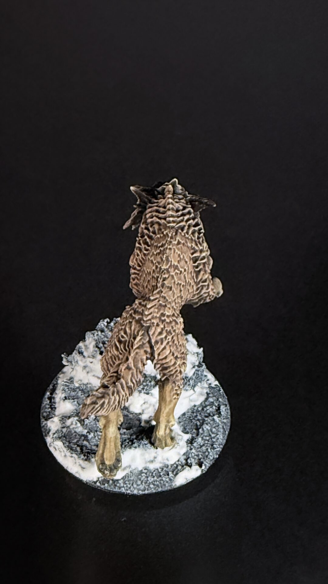Small dinosaur model figure standing on a snowy rocky base against a black background, viewed from behind.
