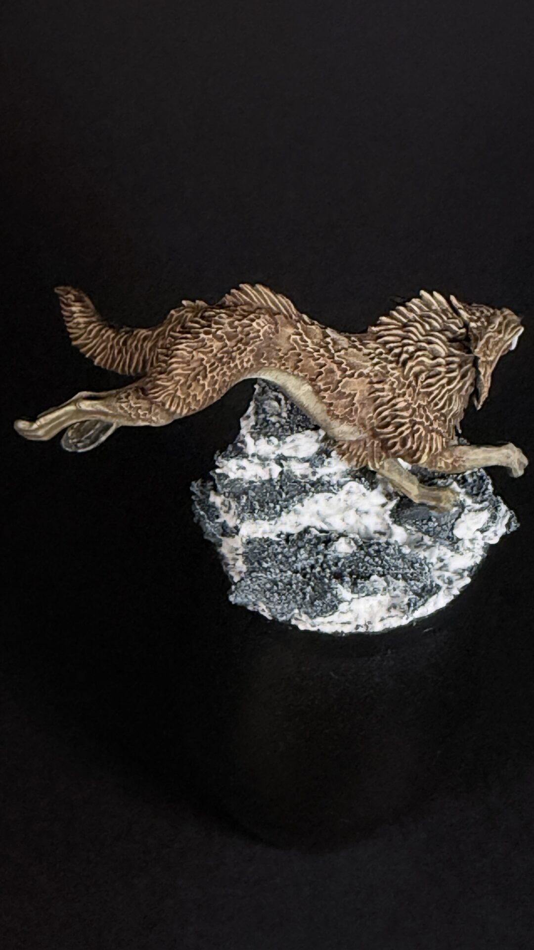 Detailed sculpture of a brown bird of prey mid-dive, perched on a snow-covered rock base against a black background.