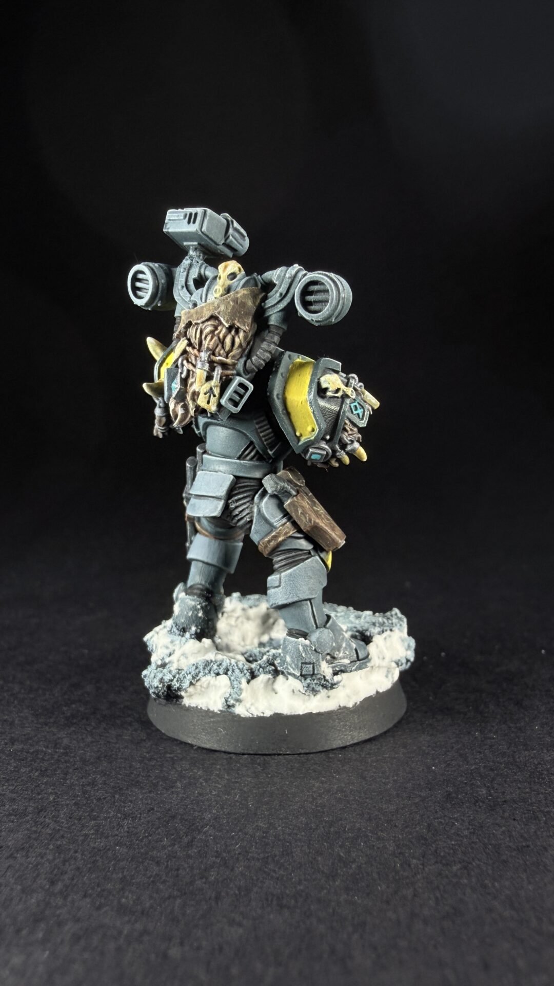 Armored sci-fi miniature figure standing on a snowy base, with a skull motif and yellow accents, wielding a large shoulder-mounted weapon system behind its back.