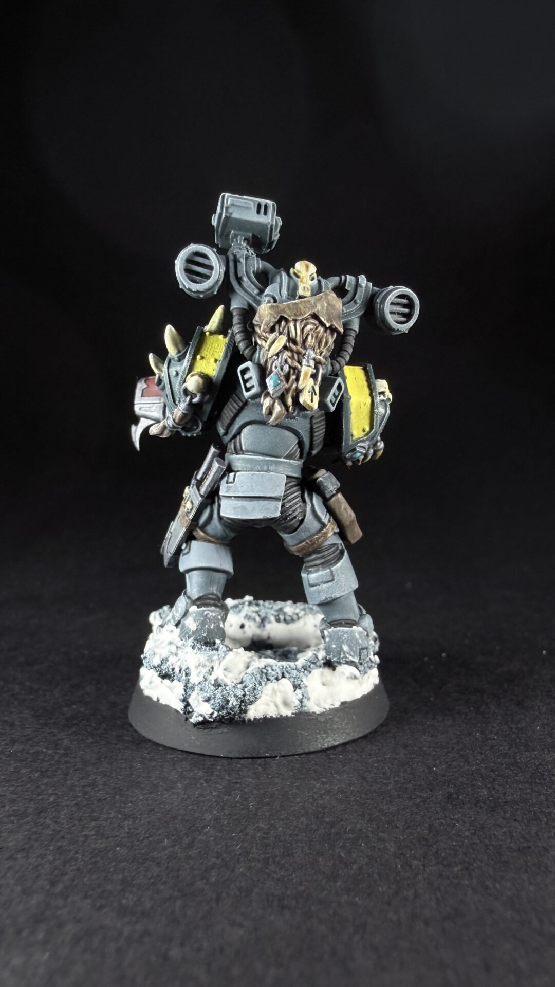 Back view of a gray armored sci‑fi figure with a large backpack, skull‑faced helm, yellow shoulder plates, standing on a snowy base.