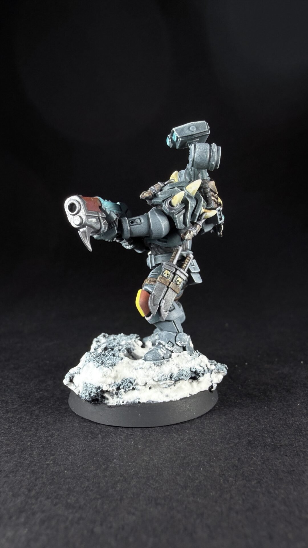 Gray armored sci‑fi mech miniature, holding a large gun, standing on a snow‑covered rocky base against a black backdrop.