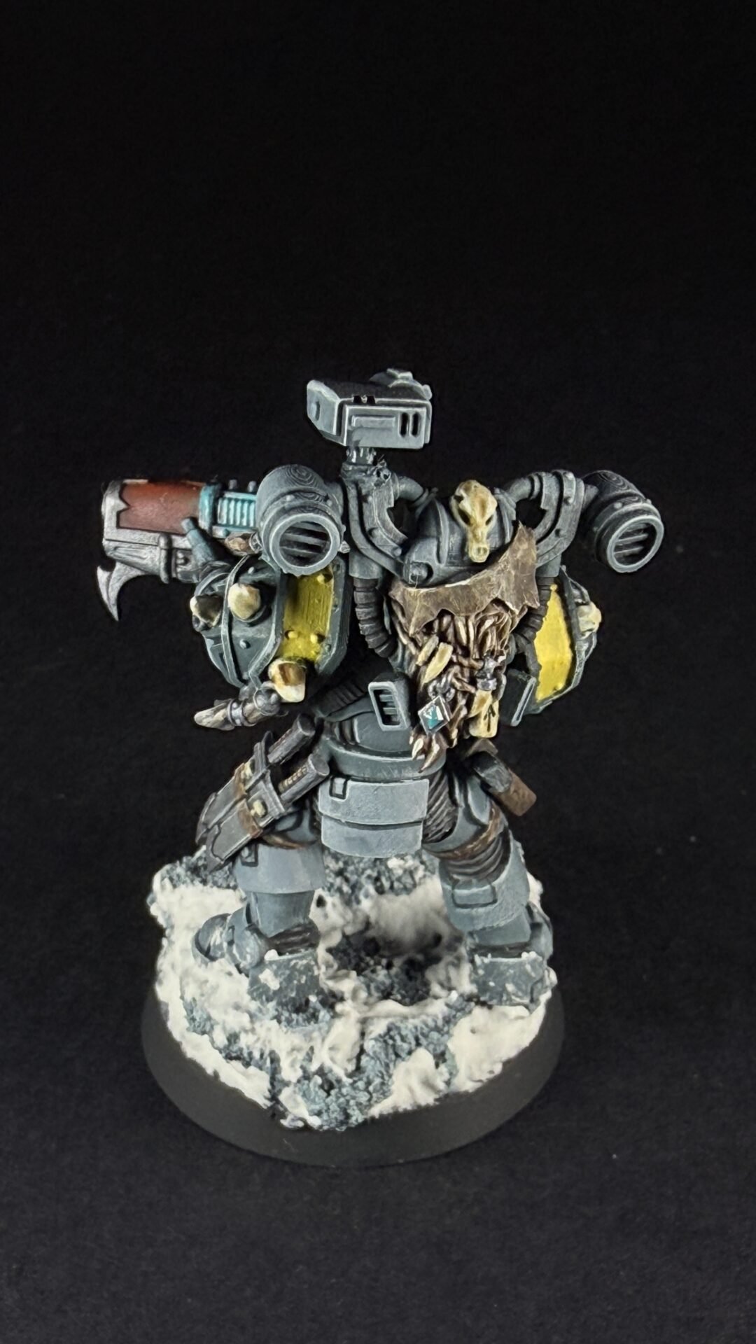 Armored sci-fi miniature figure with skulls on its chest, standing on a snowy rocky base, wielding a large weapon against a dark background.