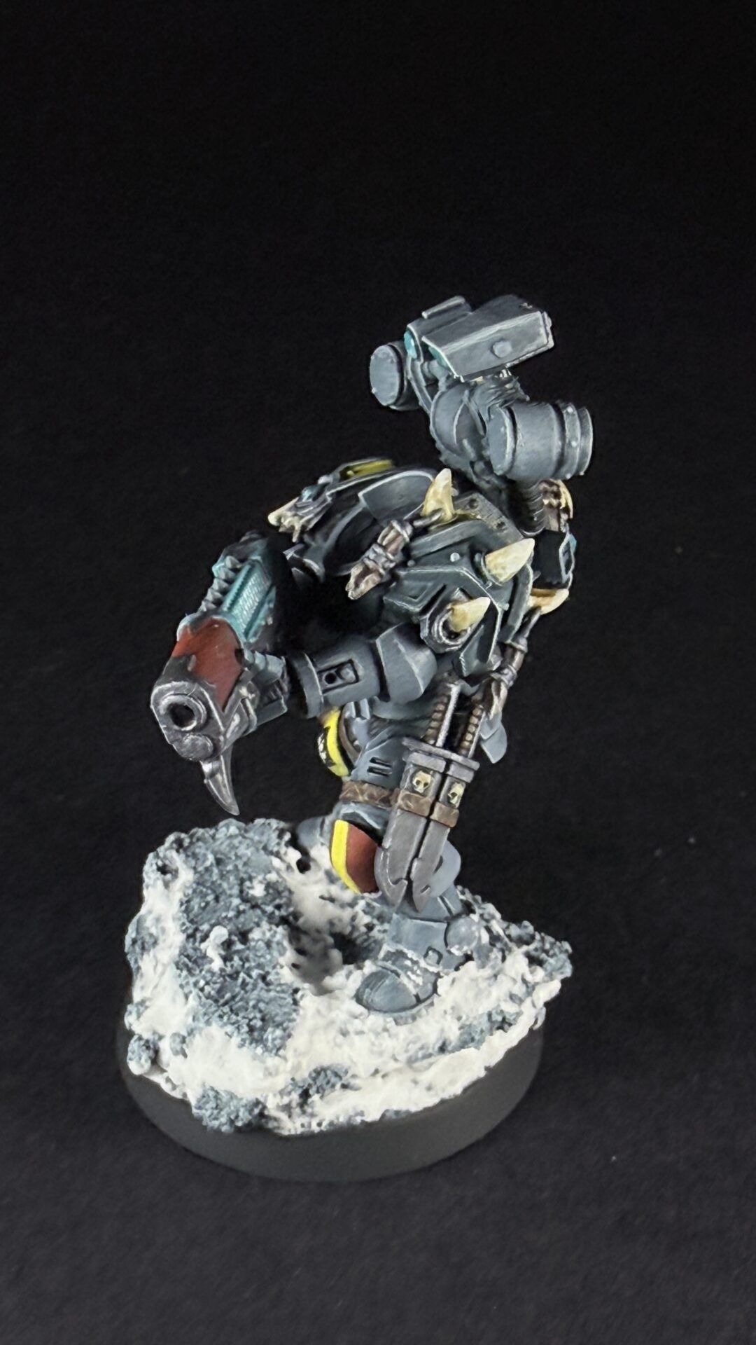 Detailed tabletop miniature: a gray, armored warrior with tusks and mechanical joints stands on a snowy rocky base, armed with a large gun on its back.