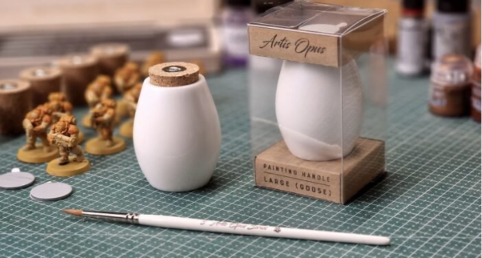 Two white ceramic vases on a green cutting mat with miniature figures and painting tools in a hobby workshop.