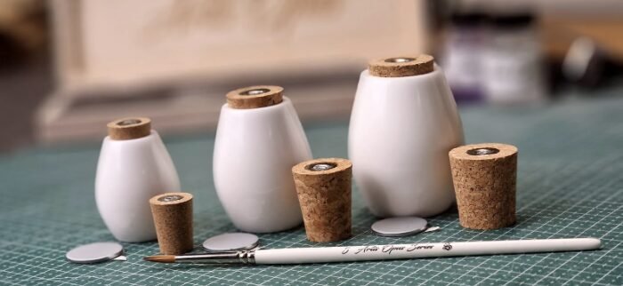 Art and craft workspace showing white ceramic bottles with cork stoppers on a green cutting mat, along with a white pencil, a craft knife, and corks.